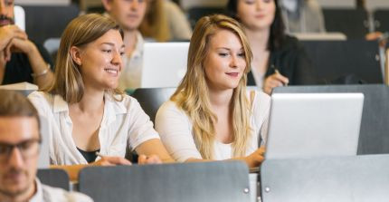 Two female students are sitting in a lecture hall with a laptop and smiling. Seated around them are other students.